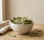 Dried bay leaves in ceramic bowl with sea salt, peppercorns and garlic on oak surface